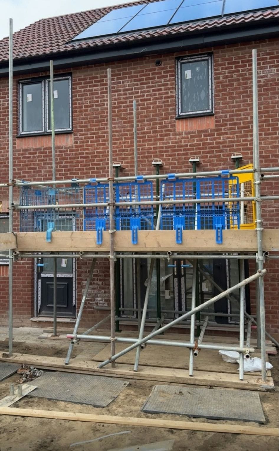 Boarded scaffold lift with blue brick guards across the front of a two storey brick house with solar panels on the roof
