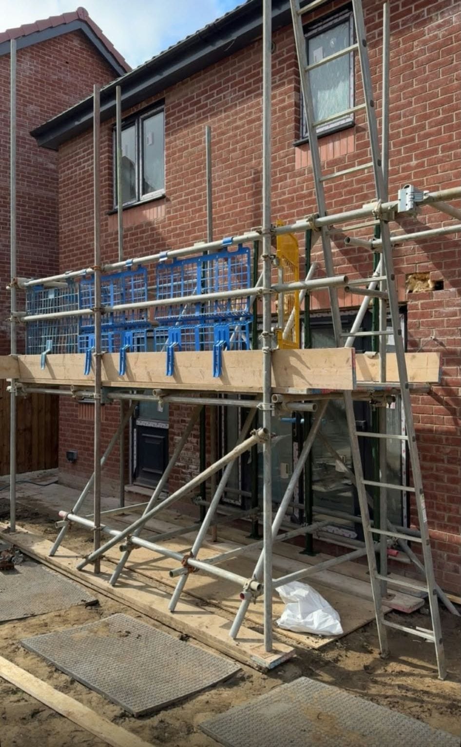 Scaffold along the back of a brick house, with a tied ladder up to a boarded lift fitted with blue brick guards