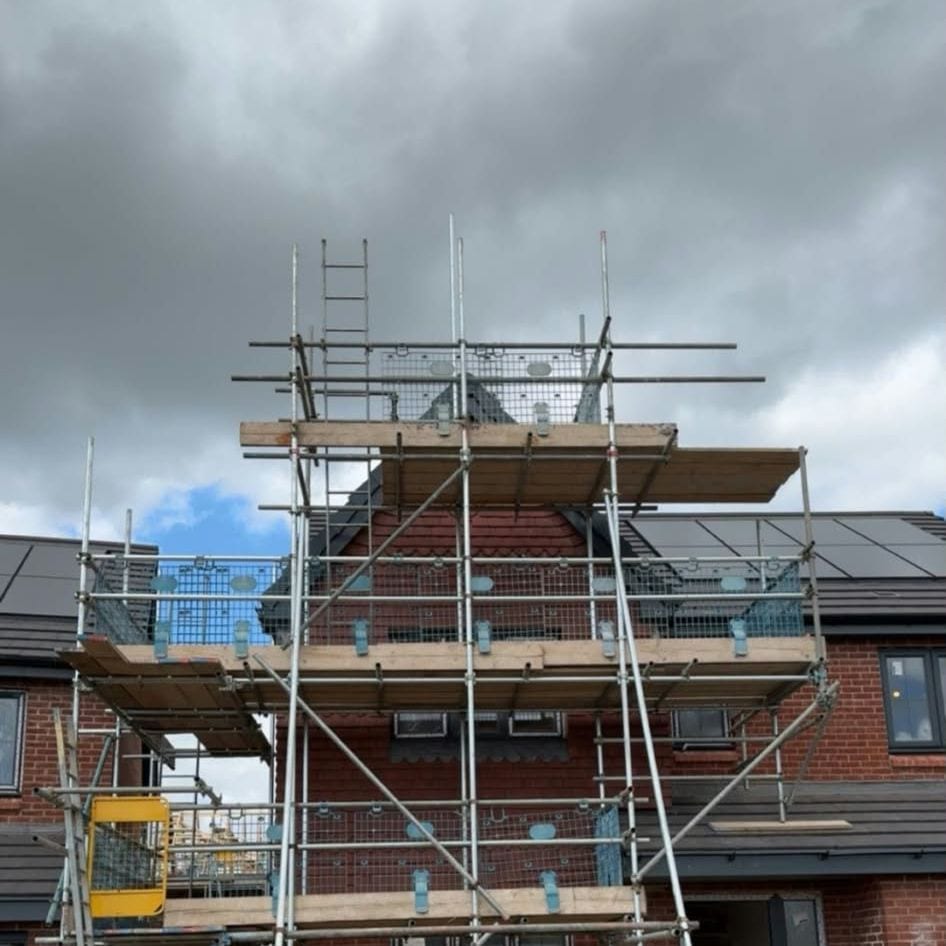 Scaffold around the gable of a new brick house, with two boarded lifts and a ladder up to the top, under a grey sky